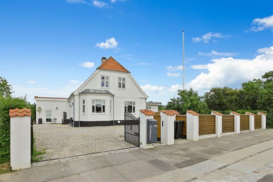 Hvidt to-etagers hus med rødt tegltag og karnap set bag en åben sort smedejernsport og grusindkørsel, flankeret af hvide piller med terracotta tagsten og træhegn, med en flagstang og klar blå himmel i baggrunden.