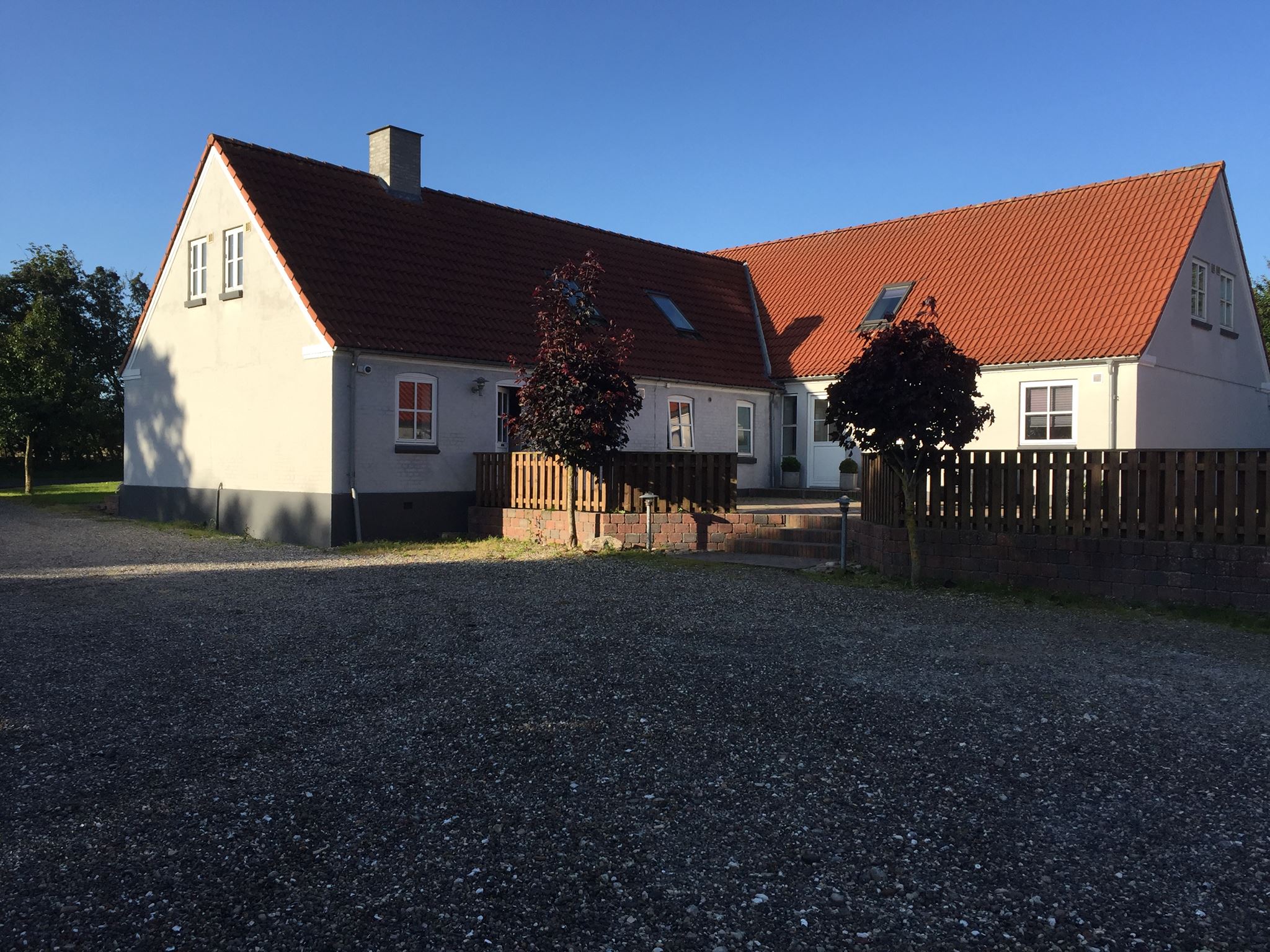 Hvidt landhus med rødt tegltag og to små purpurfarvede træer foran en lav træterrasse, omgivet af et lavt træhegn og en stor grusplads under en klar blå himmel.