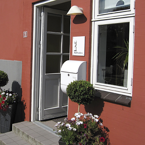 Indgang til et rødt hus med en åben hvid dør; på væggen ses en hvid postkasse, husnummeret "3" og et skilt med teksten "Hellheden" under en væglampe. Foran døren står en klippet kuglebusk og krukker med hvide og rødlige blomster ved et vindue.