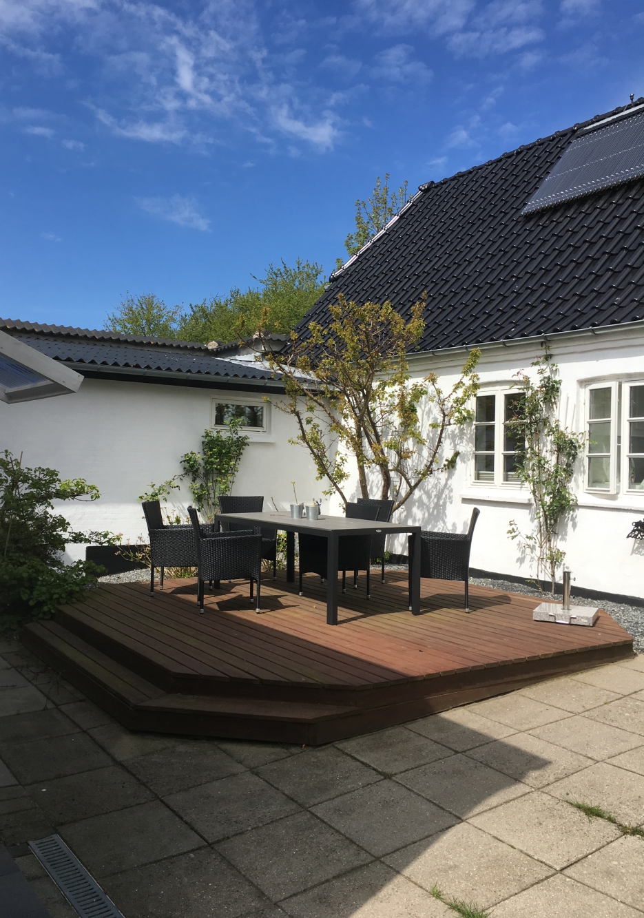 Træterrasse med et sort rektangulært havebord og fire sorte fletstole placeret midt i en lille gårdhave. Hvidt hus med sorte tagplader og et solpanel i baggrunden, en lille træbusk ved terrassen og flisebelagt gård omkring.