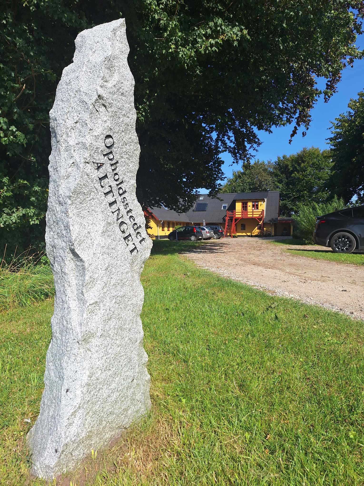En høj grå stenmonolit står på en græsplæne ved en grusindkørsel, med en gul bygning med røde trapper, parkerede biler og træer i baggrunden. Tekst på stenen: u0022Opholdsstedet ALTINGETu0022.