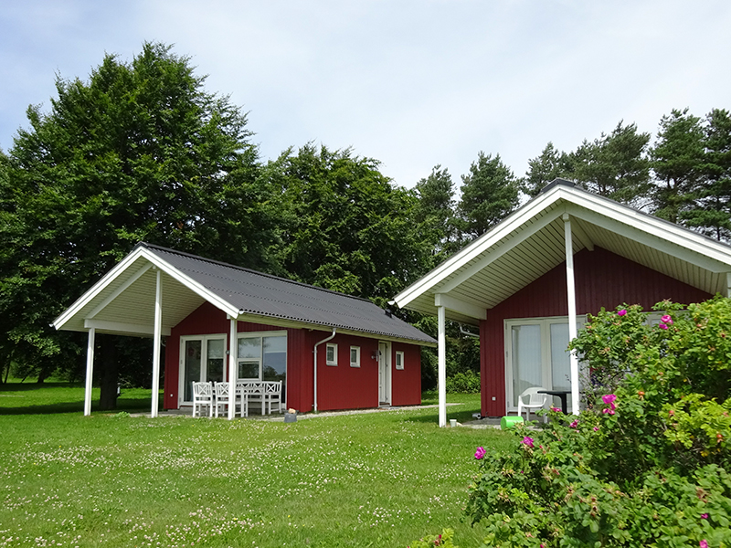 To små røde huse med hvide karme står side om side på en grøn plæne foran høje træer; det venstre har en overdækket terrasse med hvidt bord og stole, og en busk med lyserøde blomster fylder forgrunden til højre.