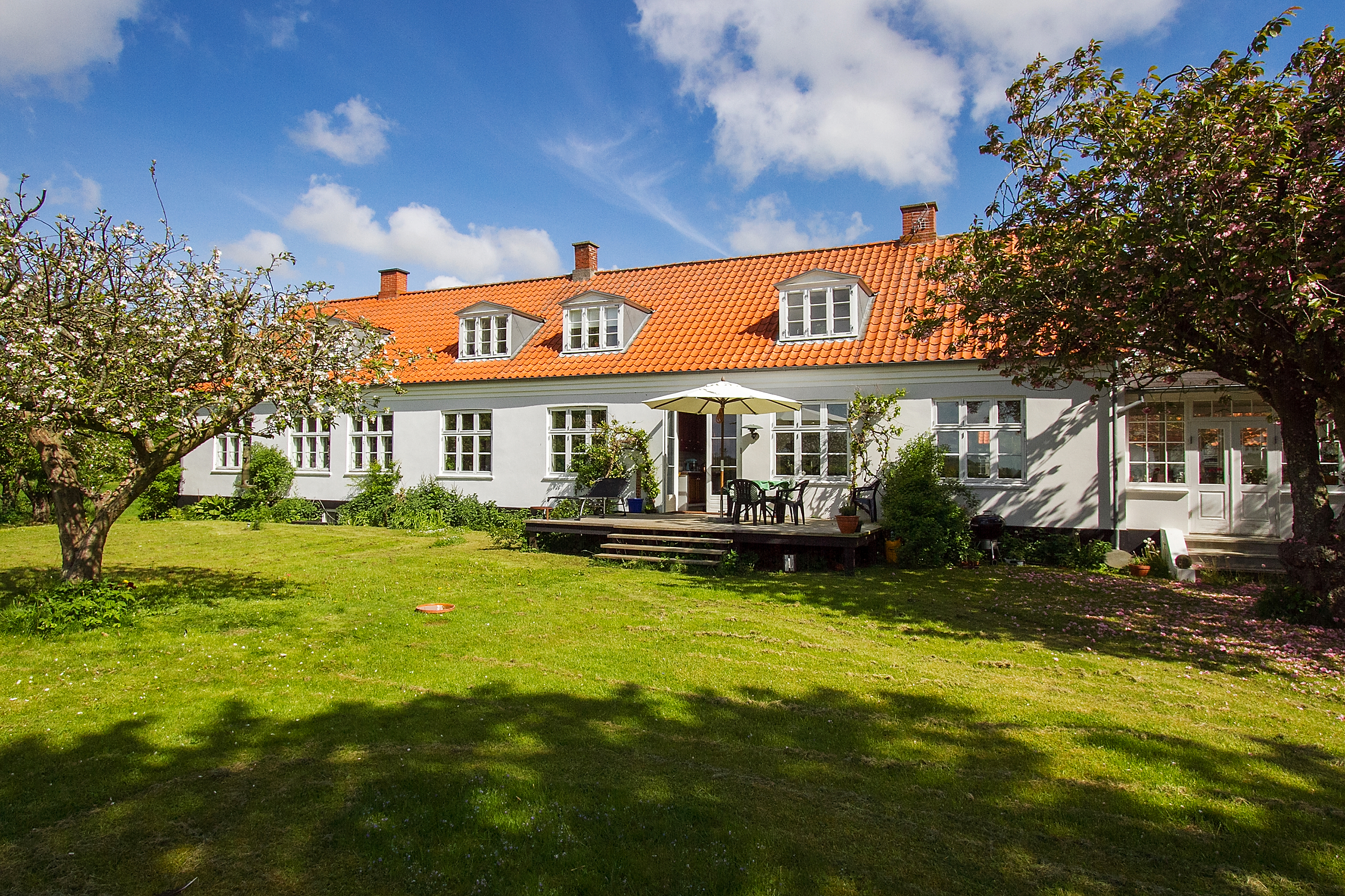 Langt hvidt hus med orange tegltag og tre kvistvinduer, terrasse med parasol og havebord foran huset; omgivet af en stor grøn græsplæne og blomstrende frugttræer under en solrig blå himmel.