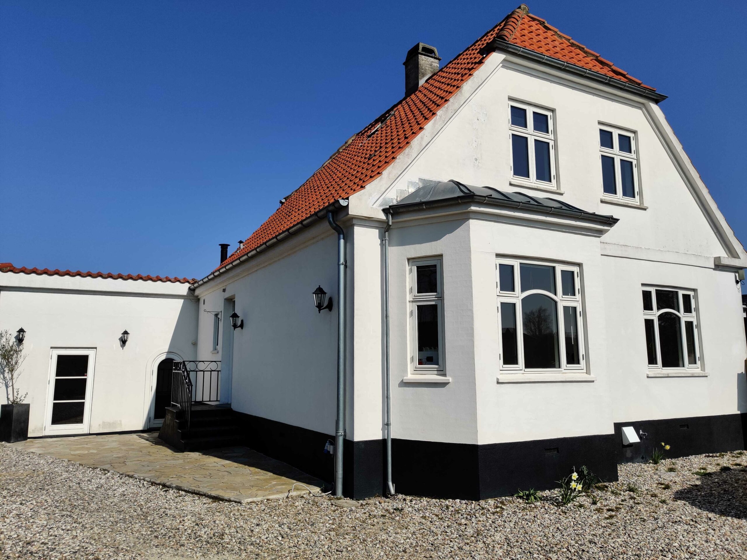 Hvidt to-etagers hus med rødt tegltag og sort sokkel, flere hvide sprossede vinduer og en lille trappeindgang i solskin. Foran huset ligger en grusbelagt indkørsel og få små blomster under en klar blå himmel.