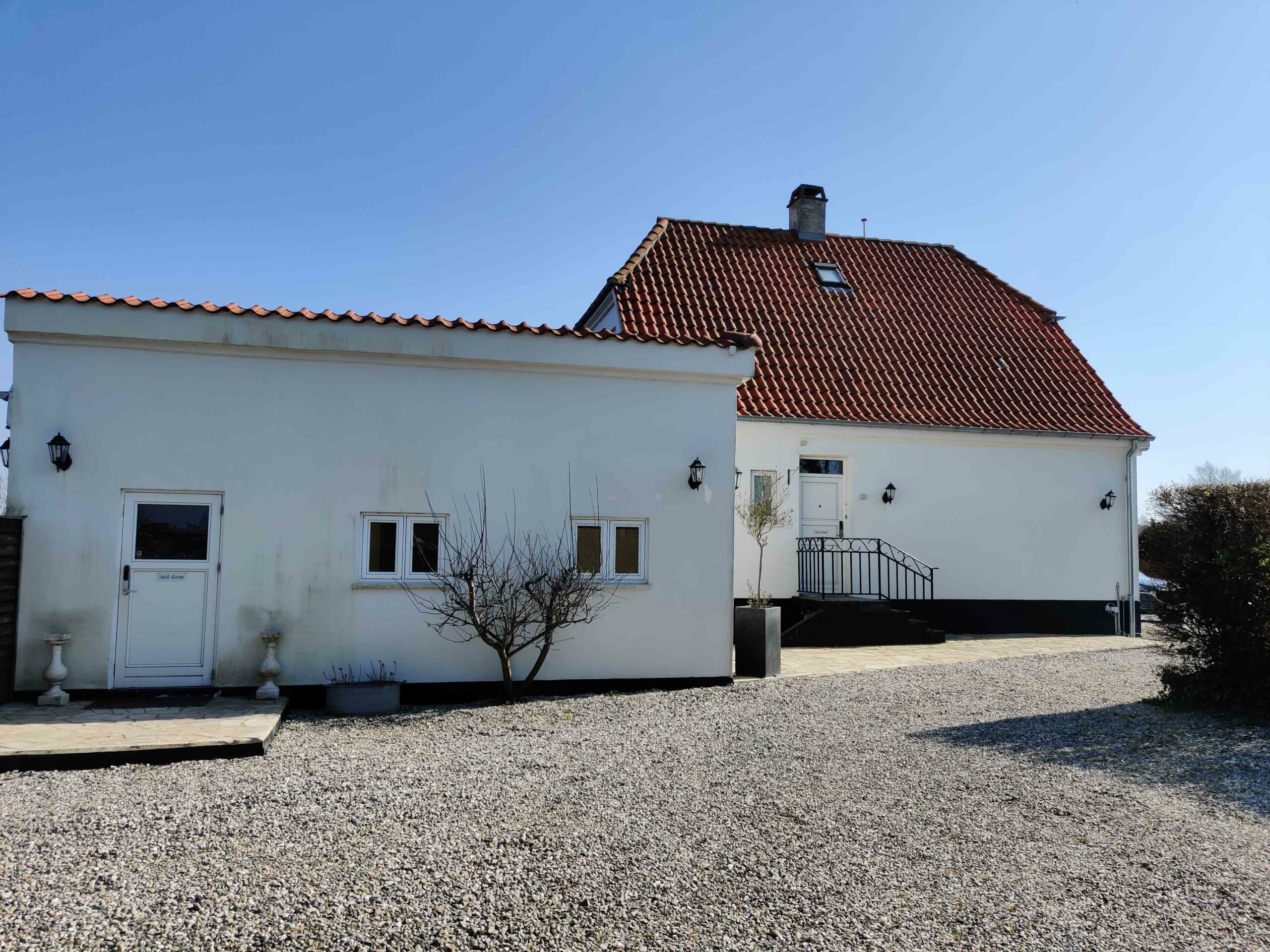 Hvidt hus med rødt tegltag står bag en stor grusbelagt indkørsel i klart vejr; en lille nøgen busk og to krukker står ved facaden, og en sort smedejernstrappe fører op til husets hoveddør.