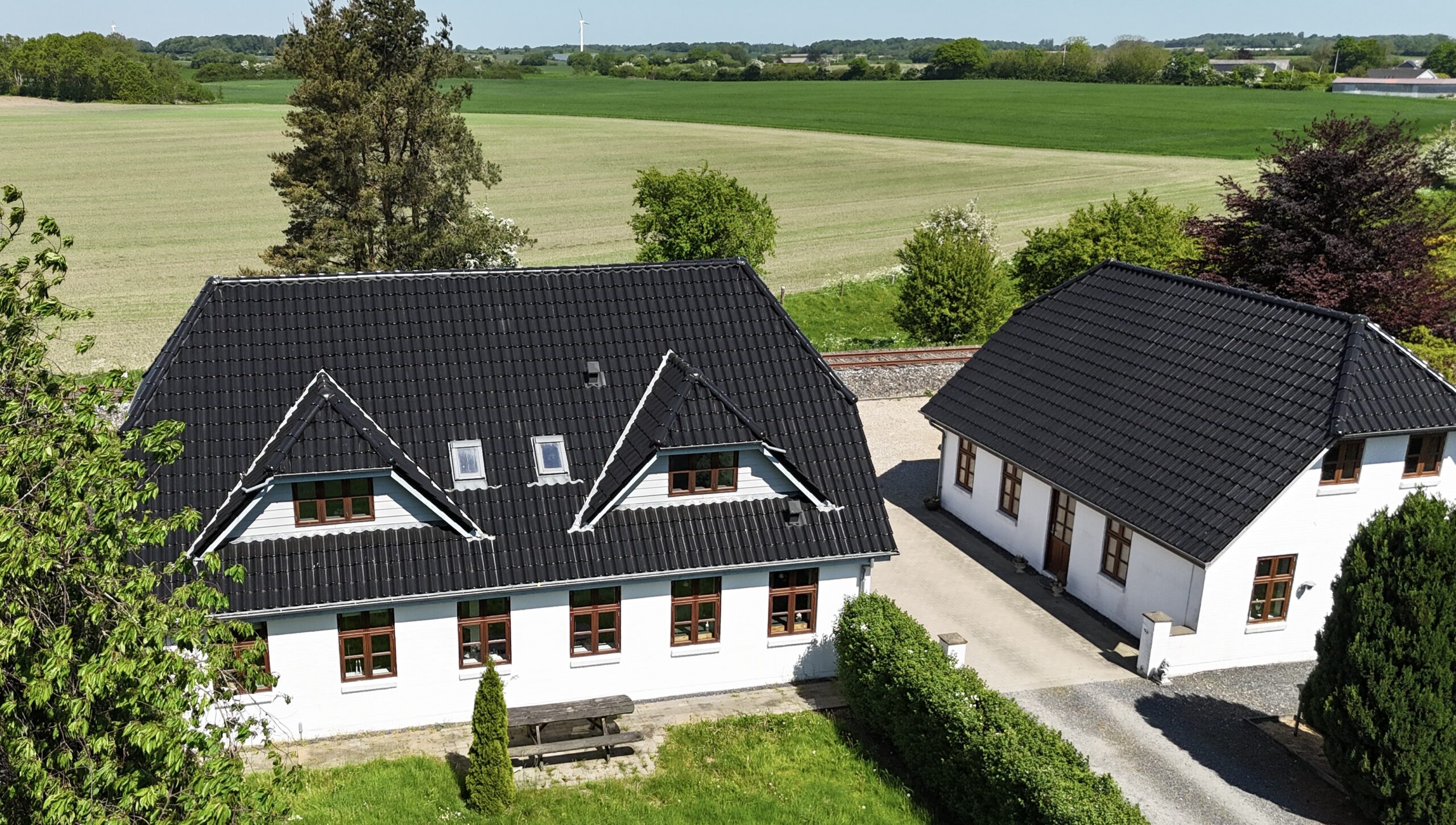 To hvidmalede gårdbygninger med sorte, spidse tagplader set skråt oppefra; den ene har et picnicbord foran, en grusgang mellem bygningerne og brede grønne marker og træer i baggrunden.