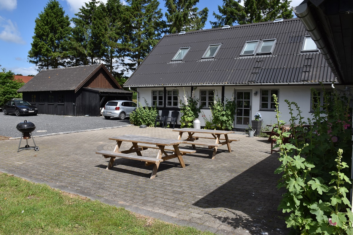 Gårdsplads med to træ-picnicborde placeret på en brostensbelagt terrasse foran et hvidt hus med mørkt skråtag og tagvinduer; til venstre ses et sort træskur og parkerede biler, og en lille kulgrill står på terrassen, mens høje stokroser vokser langs husfacaden.