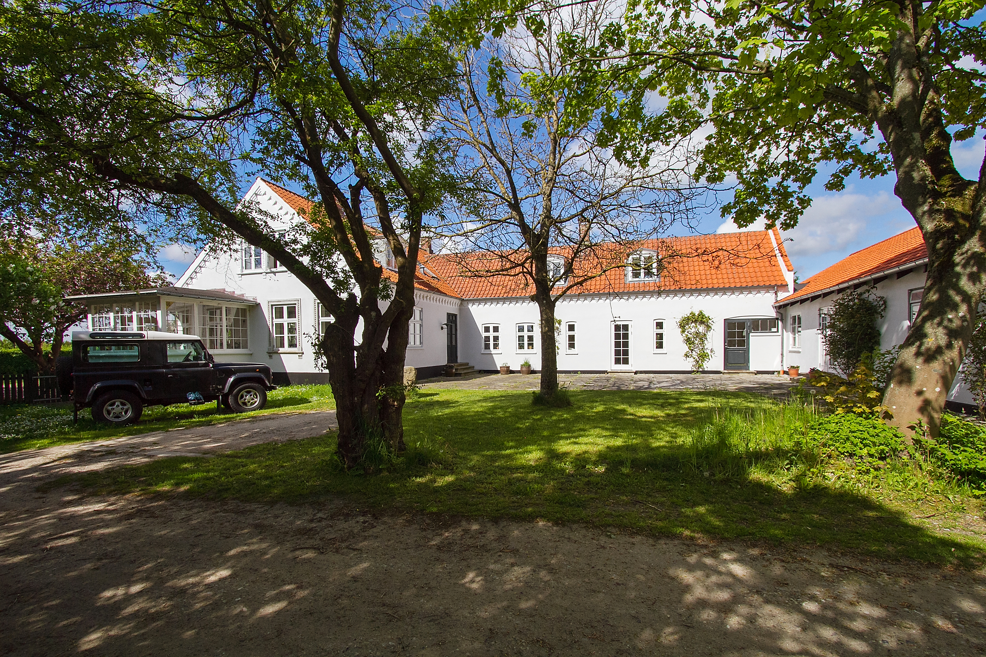 Hvid landsbygård med rødt tegltag set gennem skyggefulde træer, en mørk SUV parkeret til venstre og en grøn græsplæne og stenbelagt gårdsplads i forgrunden under en klar blå himmel.