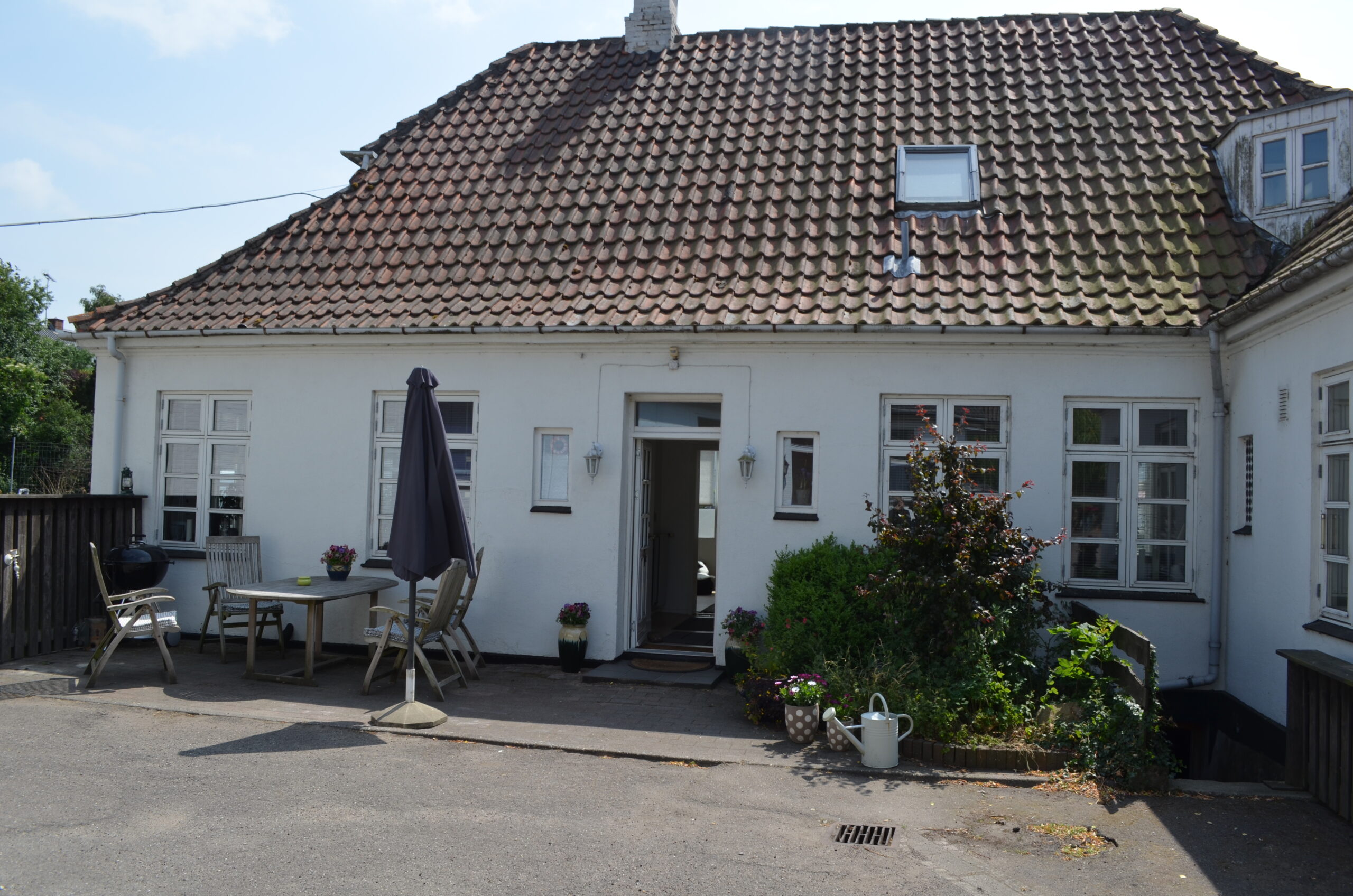 Hvidt etplans hus med rødt tegltag og en åben hoveddør, set fra en asfalteret forplads; et havebord med træstole og en sammenfoldet grå parasol står til venstre, mens krukker med blomster, buske og en hvid vandkande pryder højre side.