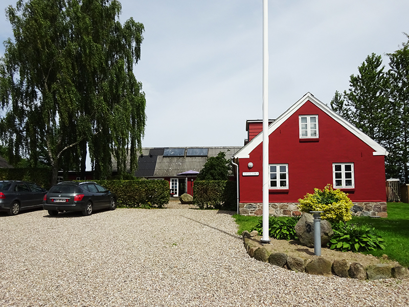 Rødt træhus med hvide vinduesrammer og stenet sokkel til højre, et hvidt flagstang foran og små bede med buske; en grusbelagt indkørsel fører mod huset med parkerede biler til venstre og høje birketræer i baggrunden.