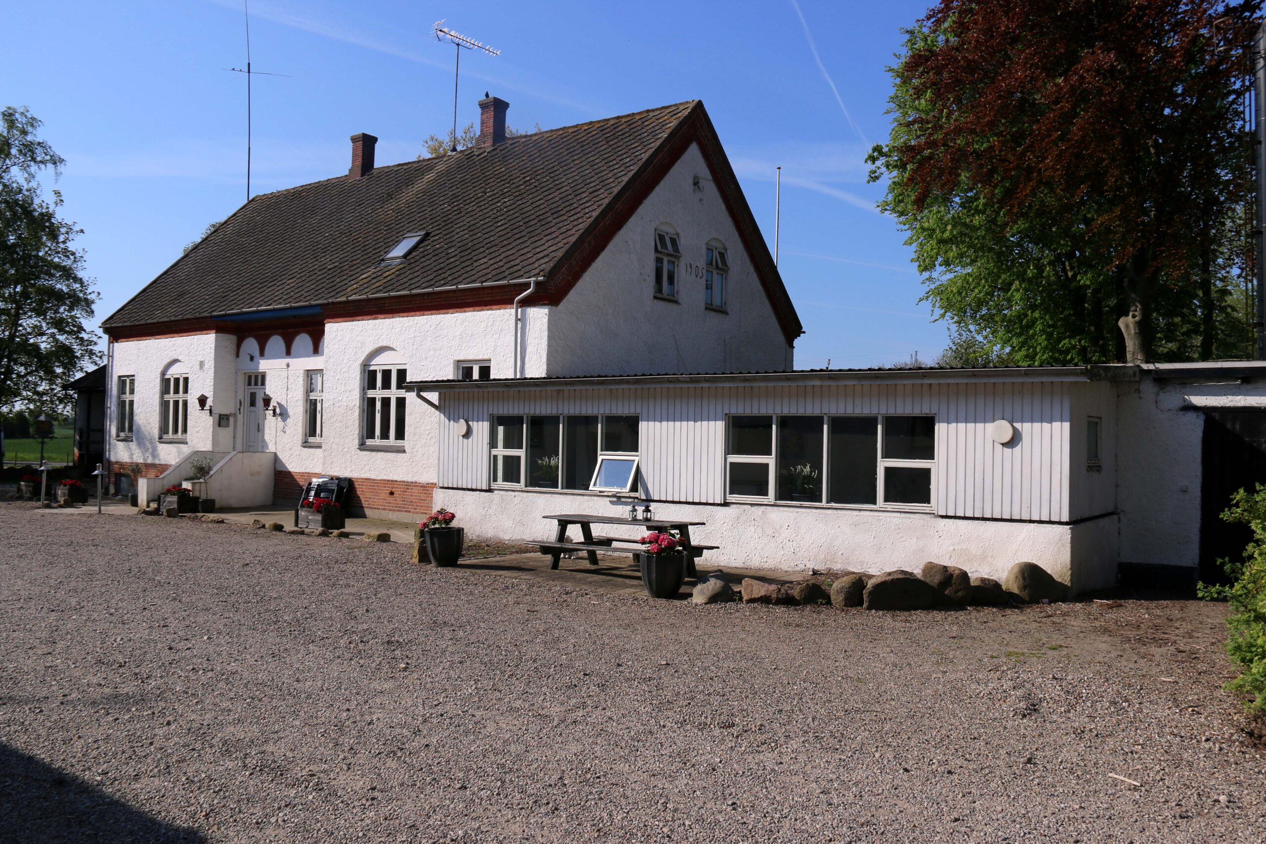 Hvidt to-etagers hus med skråt, mørkt tag og en lav tilbygning til højre står på en stor grusplads under en klar blå himmel. Foran tilbygningen står en træ-picnicbænk, flere store blomsterkrukker og en række dekorerende sten, mens træer rammer ejendommen ind i baggrunden.