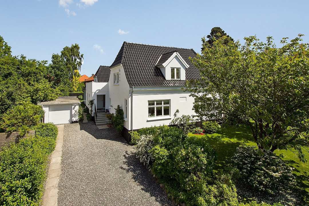 Hvidt to-etagers parcelhus med sort tag og kvistvindue ligger bag en lang grusindkørsel, der fører til en fritstående garage til venstre. Velplejet have med buske og et stort træ omgiver huset på en solrig dag under blå himmel.