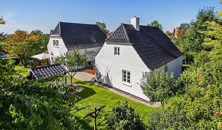 To hvide huse med mørke tagsten står i en solrig, velplejet have med grøn græsplæne, frugttræer og buske, samt en terrasse med parasol og et tørrestativ.