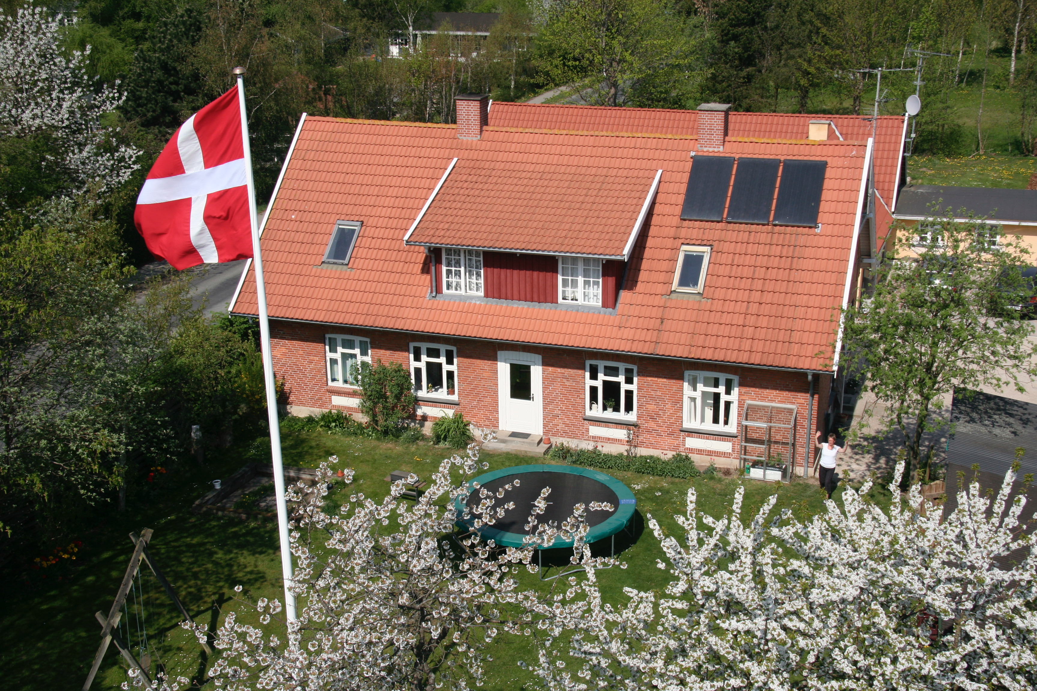 Rødt murstenshus med rødt tegltag set forfra; Dannebrog blafrer på en høj flagstang, og to solpaneler er synlige på taget. Foran huset ligger en rund trampolin på græsplænen omgivet af hvide blomstrende træer, og en person står til højre for huset med armene hævet.