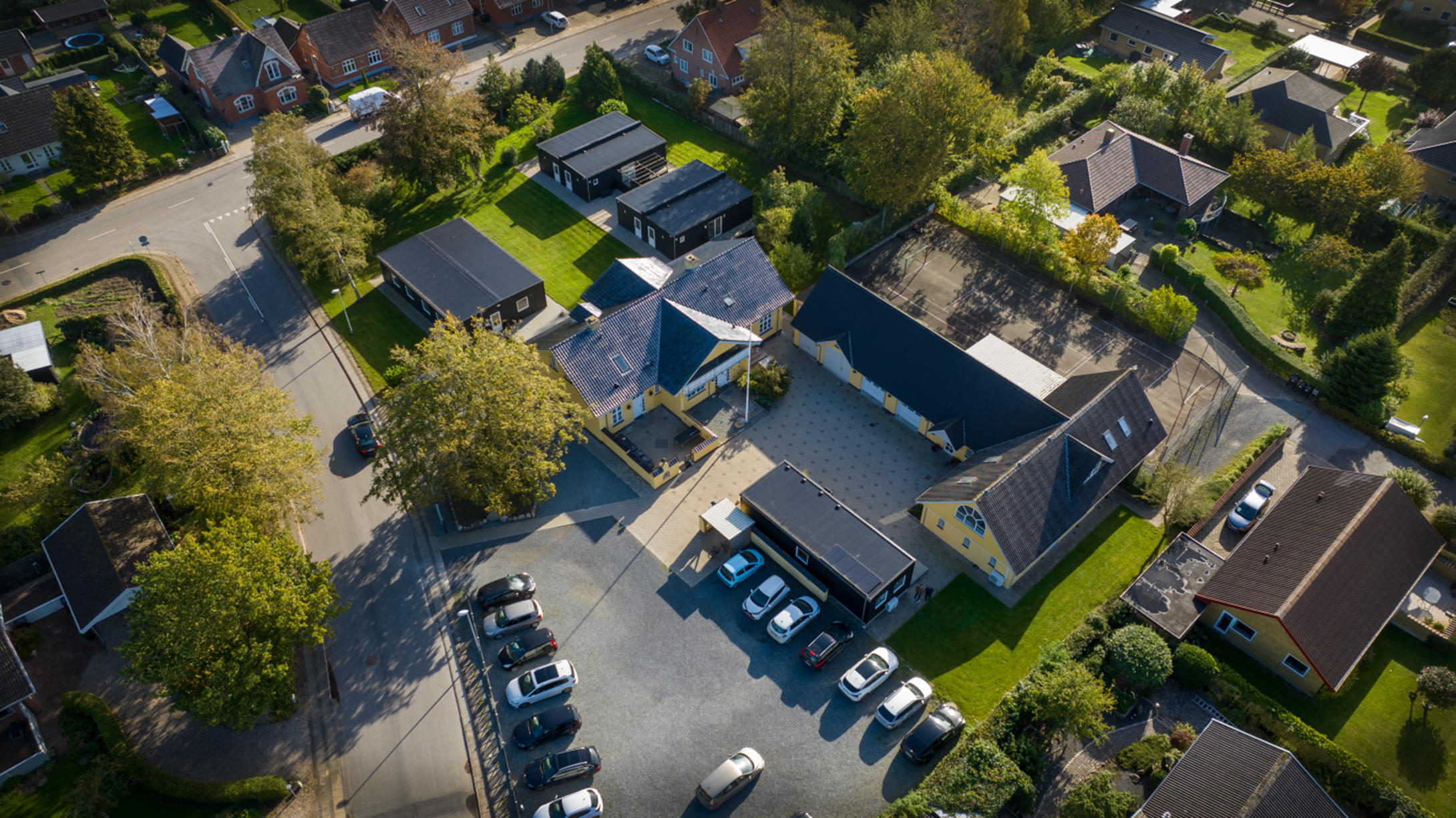 Luftfoto af en central gul bygning med en åben gårdsplads og en parkeringsplads med flere parkerede biler, flankeret af en lang sort taglænge og små sortmalede udhuse. Scenen ligger i et grønt parcelhuskvarter med træer, græsplæner og veje set ovenfra.