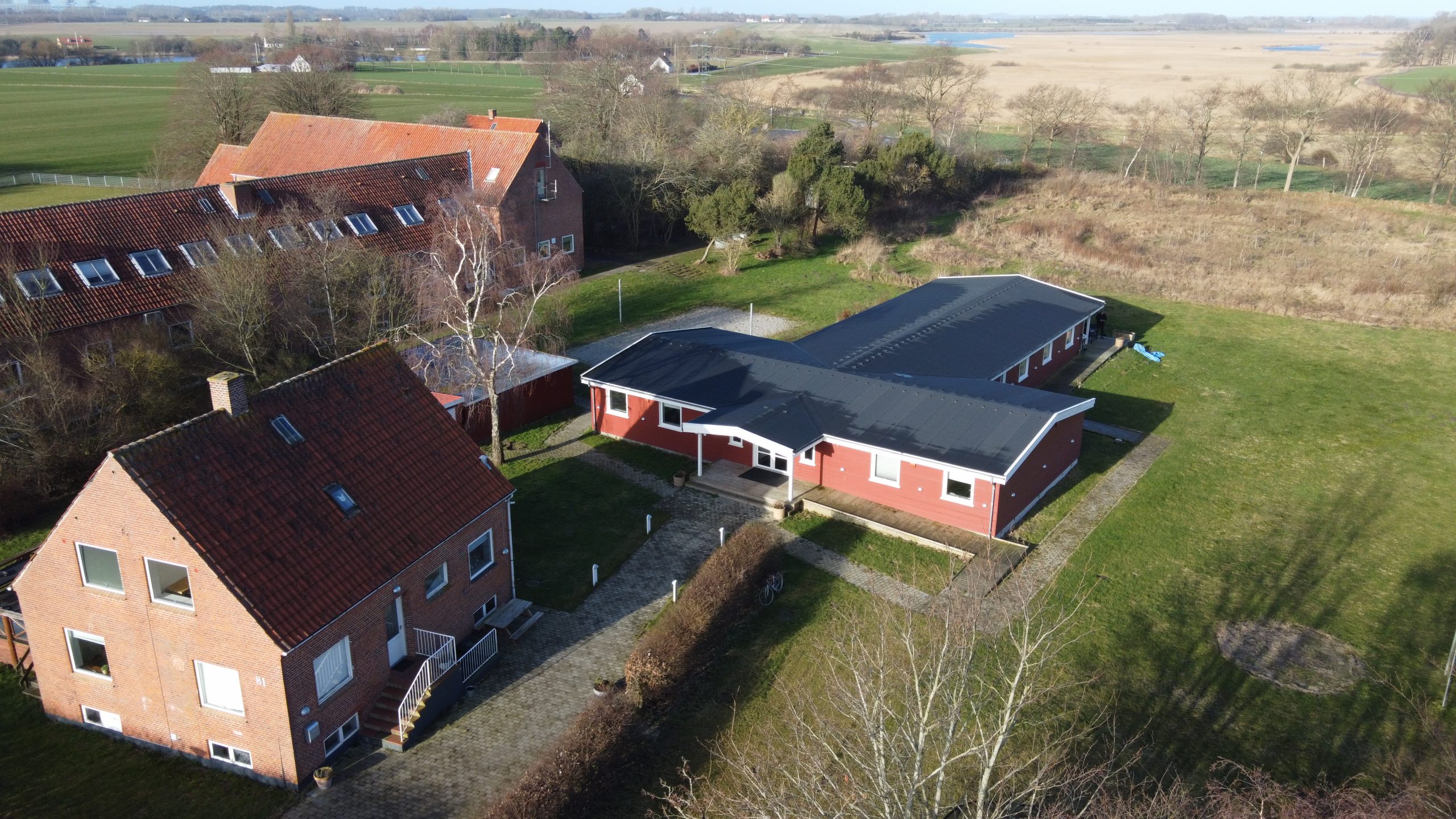 Luftfoto af to røde teglstensbygninger — et to-etagers hus med rødt tegltag forrest og en lang, lav enetages bygning med sort fladt tag bagved — forbundet af brostensstier og en lav hæk. Omkring bygningerne ligger græsplæner, enkelte birketræer og åbne marker, der strækker sig mod horisonten.