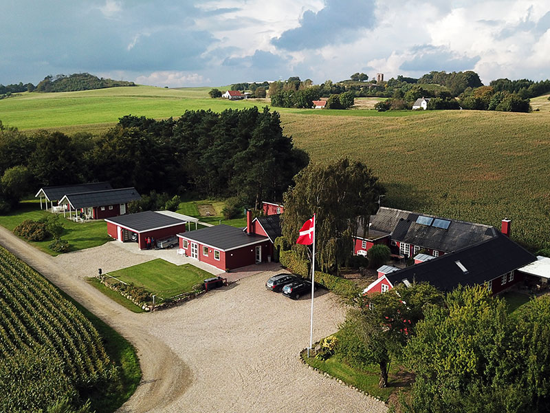 Rødt landbrugskompleks med flere lave huse og garager omkring en grusplads, to parkerede biler og et Dannebrogsflag vajende på en flagstang i forgrunden. Omgivet af marker, træer og bølgende bakker under en delvist skyet himmel.