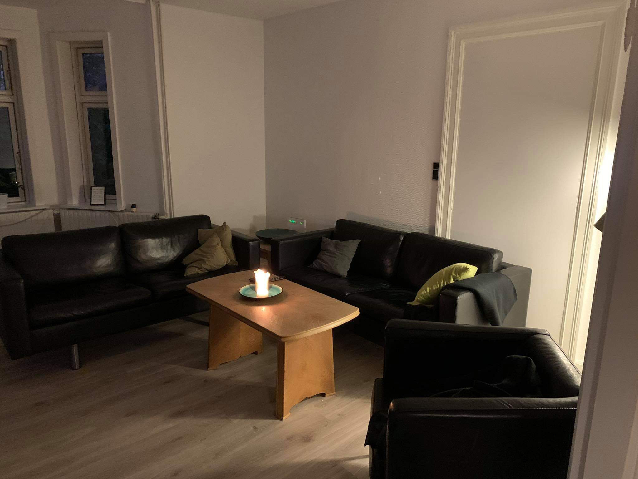 Sort lædersofaer og en sort læderlænestol står omkring et lavt træ-sofabord, hvor et tændt stearinlys på en blå fad kaster varmt lys i en ellers dæmpet stue.