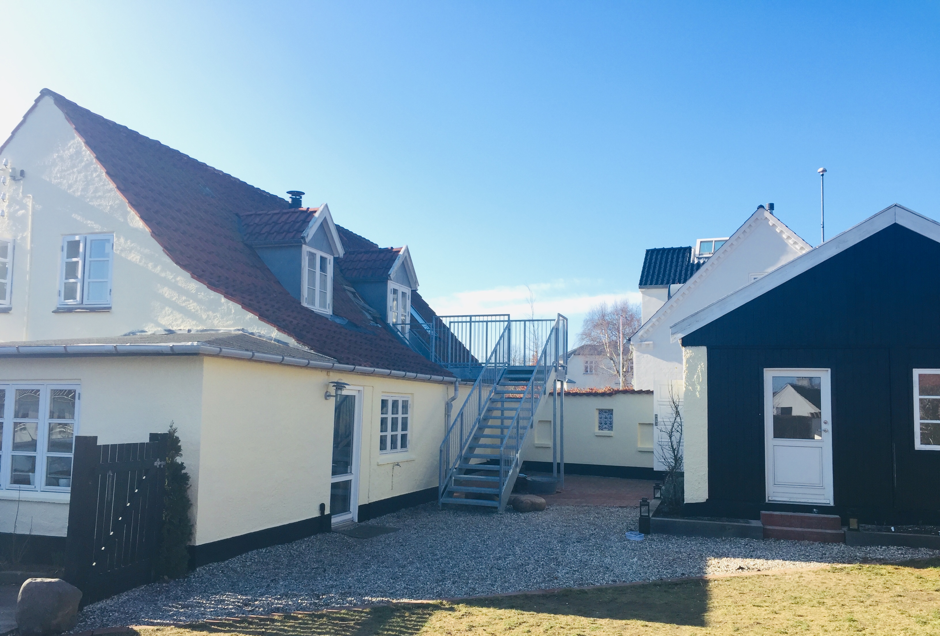 Gårdsplads med et lysegult hus til venstre med rødt tegltag og kvistvinduer, en udvendig metaltrappe der fører op til 1. sal, og et lille mørkt træhus til højre med hvid dør; grusbelagt gård og klar blå himmel.