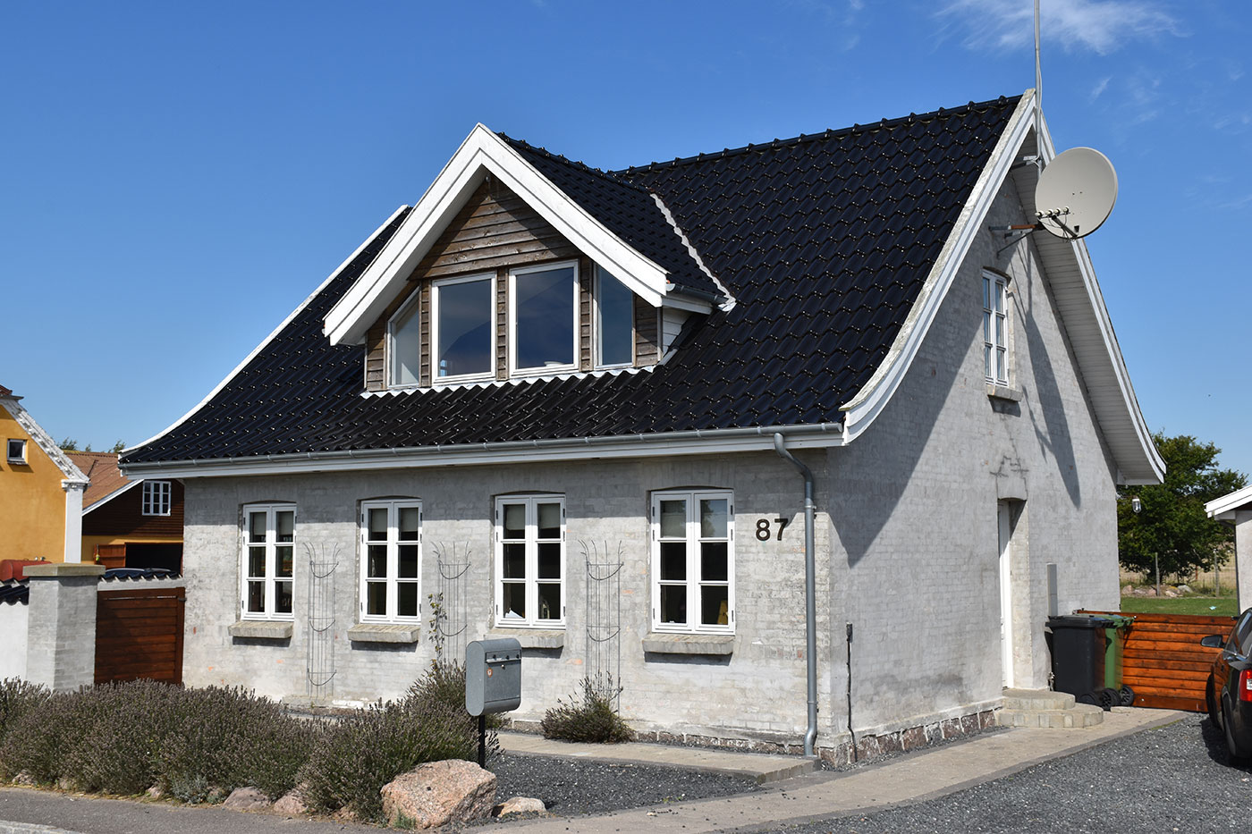 Lysegråt pudset parcelhus med sort, glinsende tag og en kvist med tre vinduer, hvide rammer, satelliteskål på gavlen og husnummeret 87 synligt ved facaden; foran er lave buske, en postkasse og en grusbelagt indkørsel.