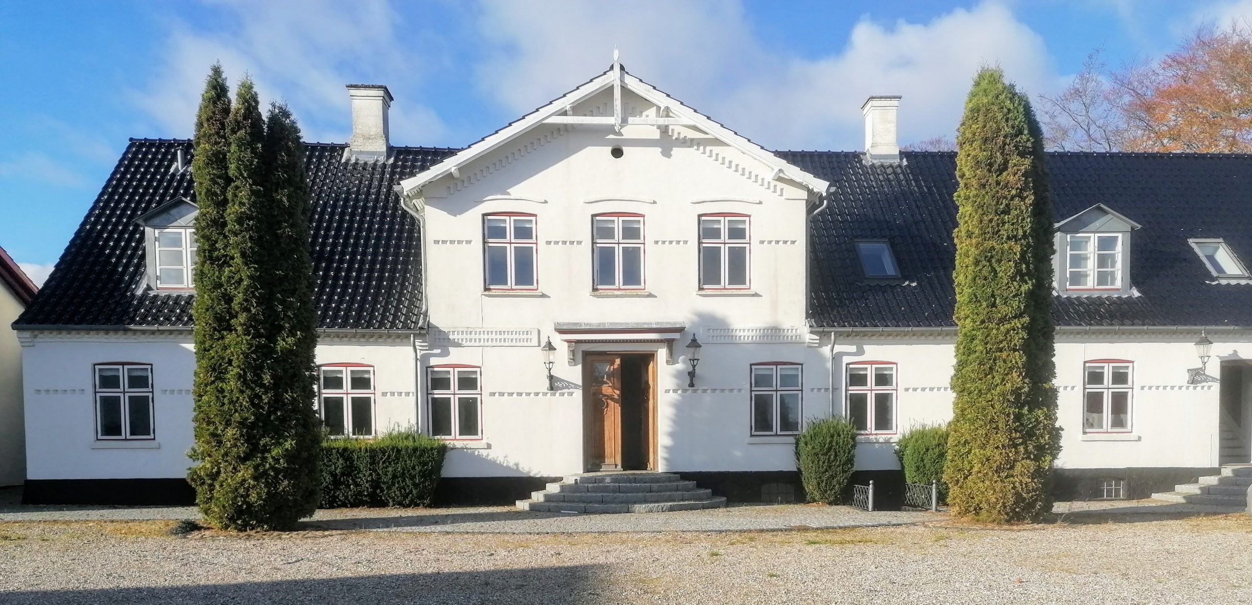 Hvidt, symmetrisk to-etagers hus med sort tegltag og en midterstillet trædør på en rundet trappesats; facade har flere sprossevinduer og tagkvist. To høje, slanke stedsegrønne træer står symmetrisk i forgrunden på en grusbelagt indkørsel.
