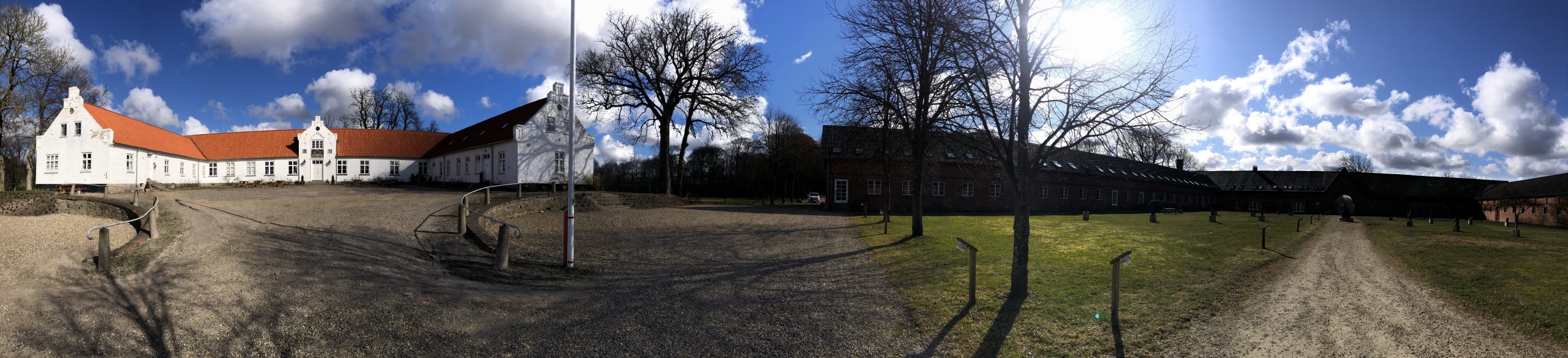 Hvid, L-formet hovedbygning med rødt tegltag til venstre og en lang rød murstenslænge til højre, adskilt af en stor grusgård med en central sti, en flagstang og nøgne træer under en blå himmel med hvide skyer.