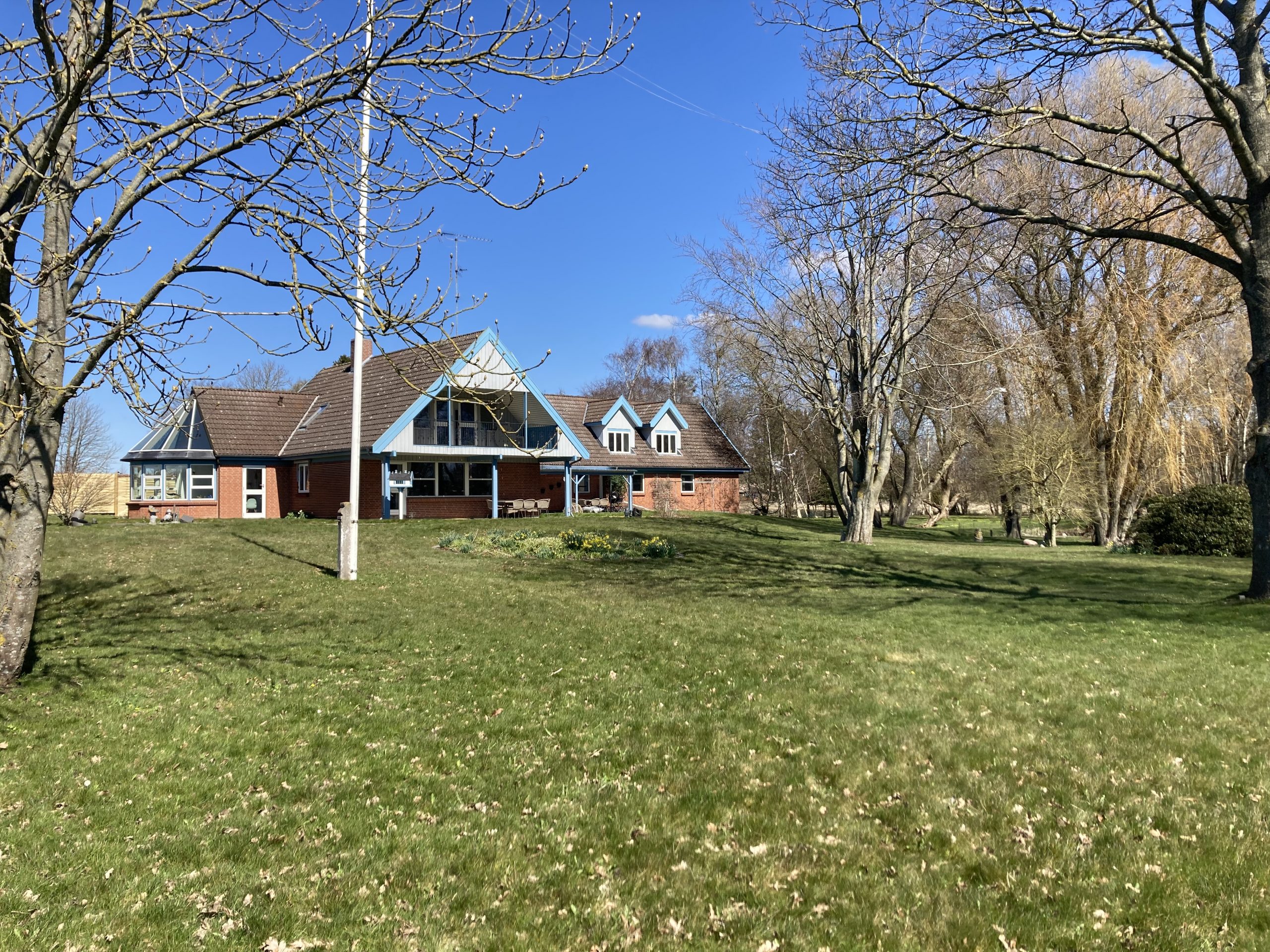 Rødmuret hus med blå gesimser og saddeltag står på en stor grøn græsplæne under en klar blå himmel; en flagstang og et blomsterbed med gule påskeliljer ses foran huset, omgivet af nøgne forårstræer.