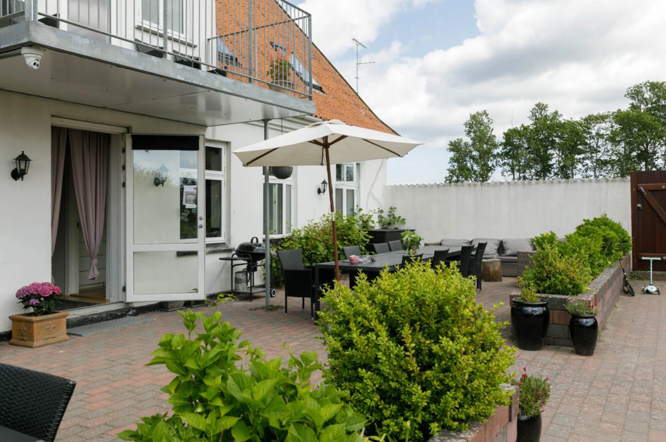 Terrasse med et langt sort havebord under en åben hvid parasol, omgivet af sorte stole og grønne buske i plantekasser på en flisebelagt gårdhave; bagved ses et hvidt hus med åben dør og lyserødt gardin, en balkon over indgangen, grill, sofahjørne og en træport i baggrunden.