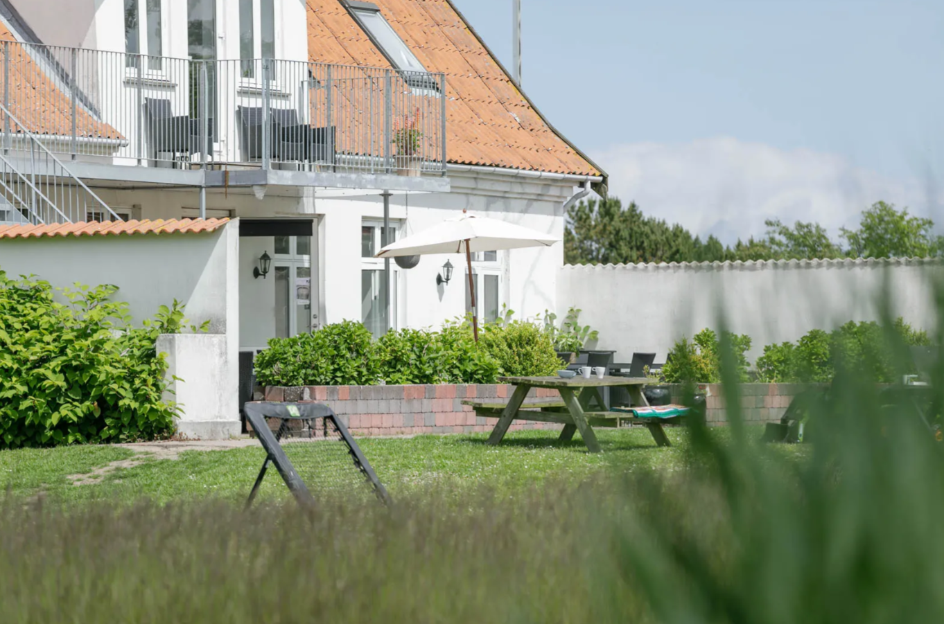 Hvidt hus med orange tegltag og metalaltan set fra en forhave; foran huset ligger en græsplæne med picnicbord, parasol, liggestole og lave blomsterbede omgivet af grønne buske.