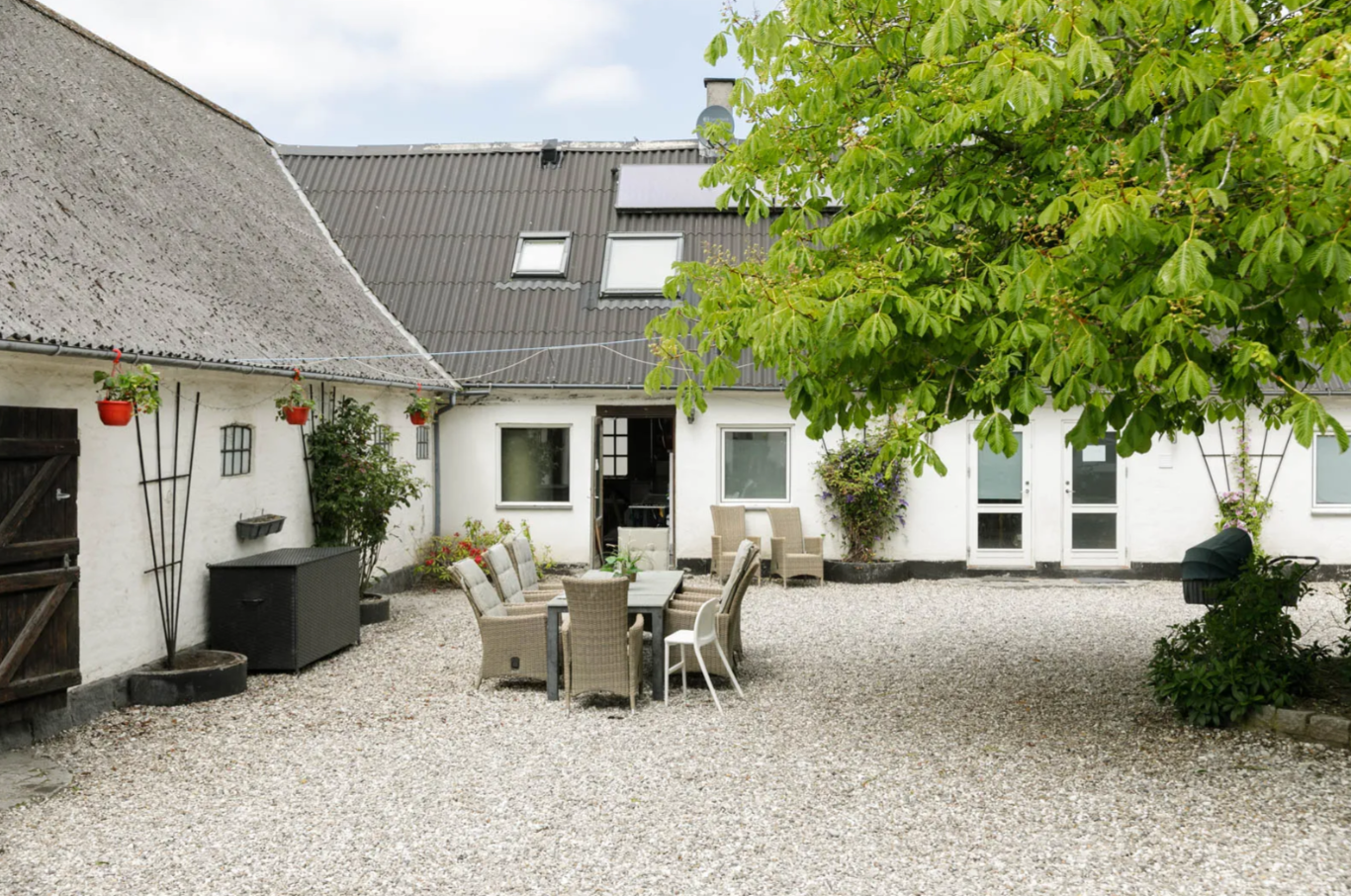Gårdsplads med hvide bygninger og grusbelægning, hvor et langt udendørs spisebord med fletstole står midt i gården. Et stort grønt træ kaster skygge til højre, hængende røde plantekrukker langs facaden og en åben dør i baggrunden.