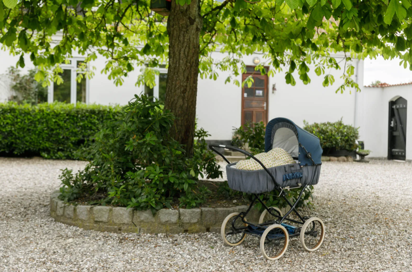 En blå retrobarnevogn står parkeret under et stort grønt træ på en gruset gårdsplads foran et hvidt hus med en trædør.