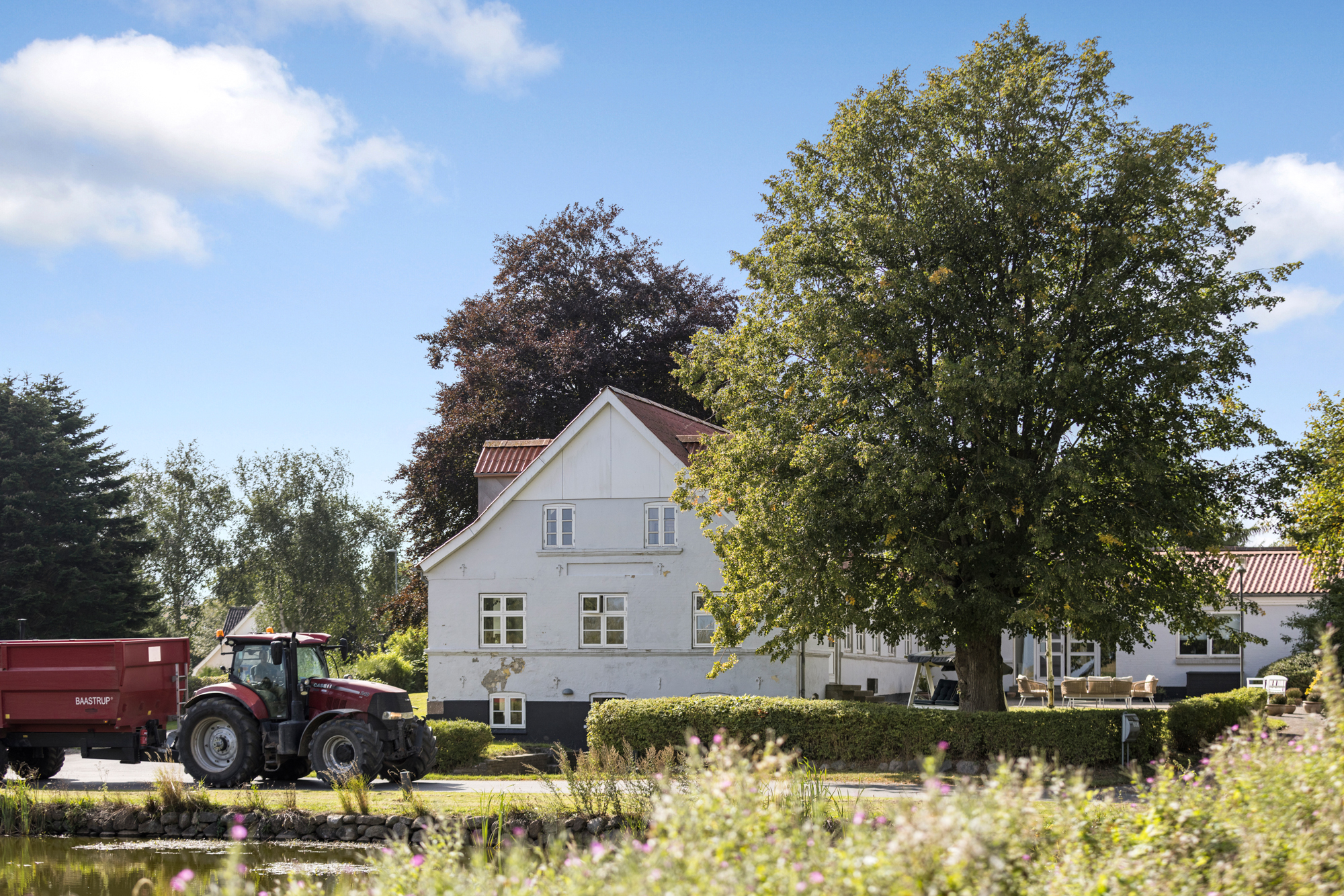 Hvidt landhus står bag et stort løvtræ, mens en rød traktor med en rød anhænger mærket u0022BAASTRUPu0022 holder langs vejen foran huset. I forgrunden ses en dam med slørede blomster og en klar blå himmel med få hvide skyer.