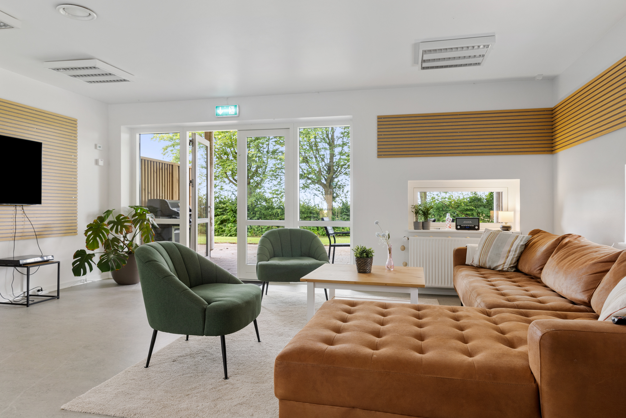 Stor brun chaiselong sofa og to grønne polstrede lænestole står omkring et lavt træbord i et lyst, moderne opholdsområde. Glasterrassedøre bagtil står åbne mod en terrasse og træer, og rummet har dekorative trælameller på væggene samt et vægmonteret tv.