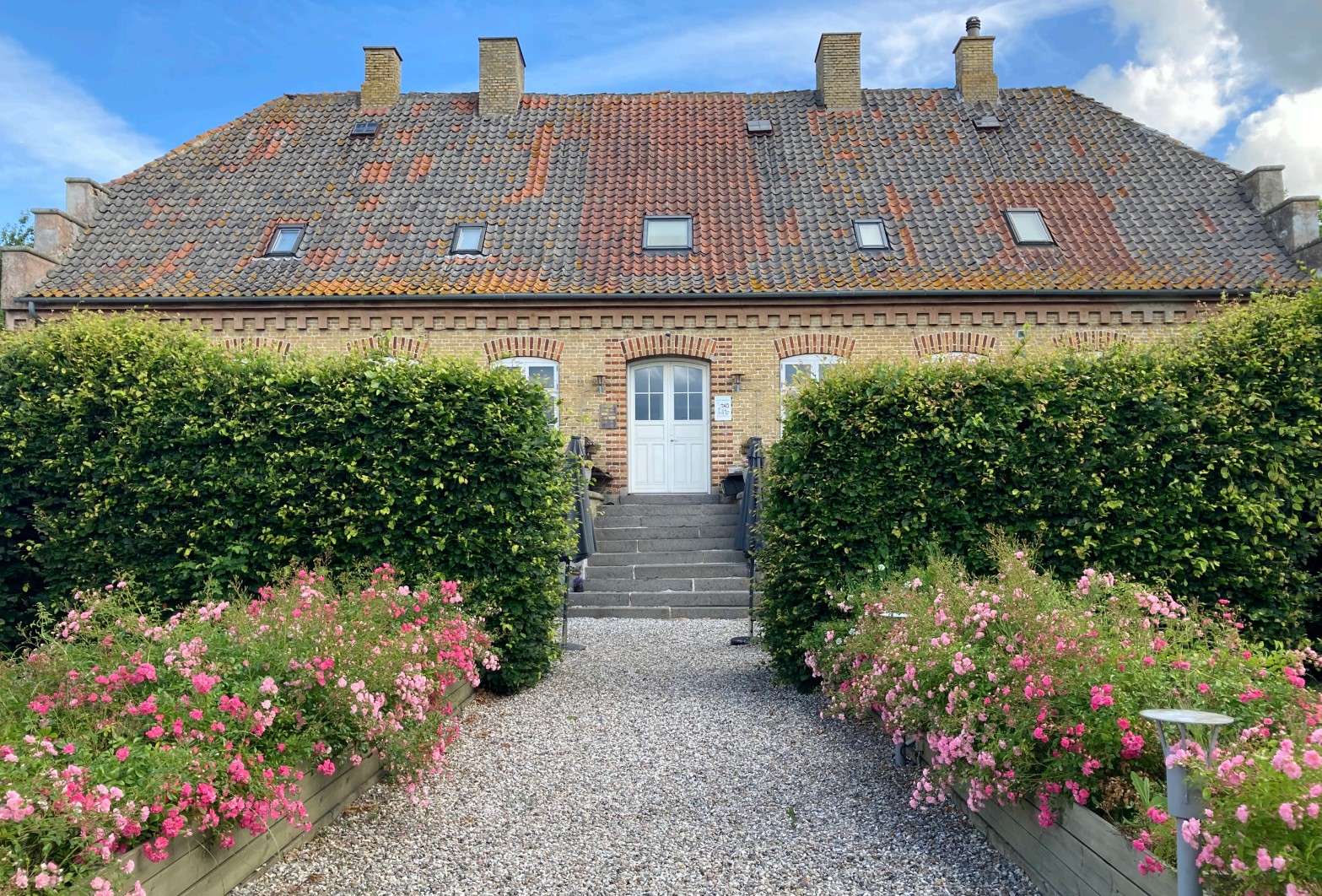 Rækkehus i gult murværk med stort rødbrunt tegltag og en hvid dobbeltdør øverst på en kort trappe midt på facaden. En grussti fører op mellem to høje, trimmede hække og plantekasser fyldt med lyserøde blomster under en blå himmel.