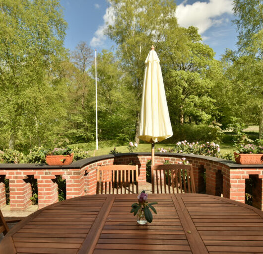 Rundt træbord med stole på en terrasse, et lille glas med en blomst står på bordet. En sammenfoldet lys parasol står i midten foran en rød murstensbalustrade med blomsterkummer og frodige grønne træer under blå himmel.