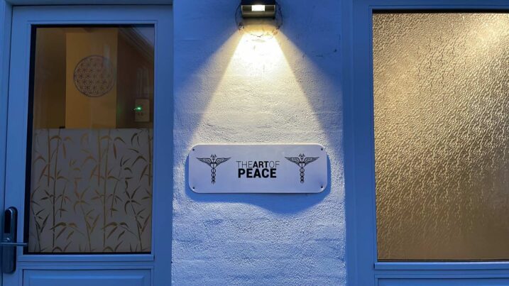 Hvidt rektangulært skilt med teksten u0022THE ART OF PEACEu0022 i sort flankeret af to kaduceus-symboler, monteret på en hvid pudset væg under en væglampe, mellem en dør med mønstret frostet glas og et større frostet vindue.