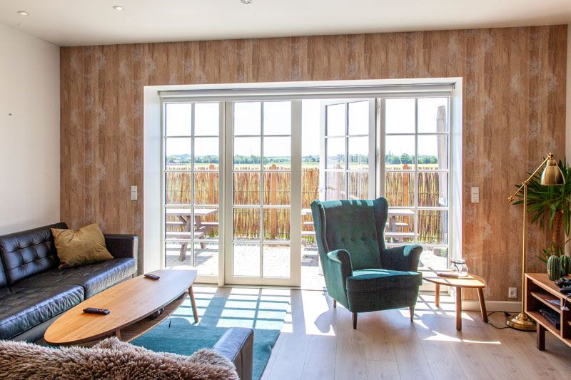Lyst opholdsstue med store franske glasdøre, der åbner til en solbeskinnet terrasse, hvor sollys danner skygger på det lyse trægulv. En grøn lænestol står ved dørene, en sort lædersofa til venstre med et ovalt træsofabord foran samt en gulvlampe og planter i højre hjørne.