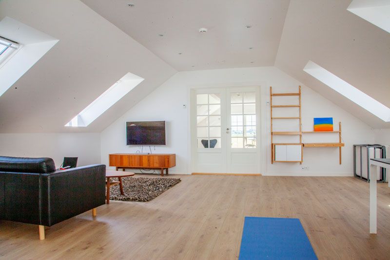 Loftstue med skråtag og indbyggede ovenlysvinduer, lyst trægulv og en blå yogamåtte i forgrunden. Til venstre står en sort lædersofa foran et vægmonteret tv over et træskab og et lille sofabord på et mørkt tæppe; i midten er hvide franske døre, og til højre hænger en træladdersreol med et lille farverigt maleri.