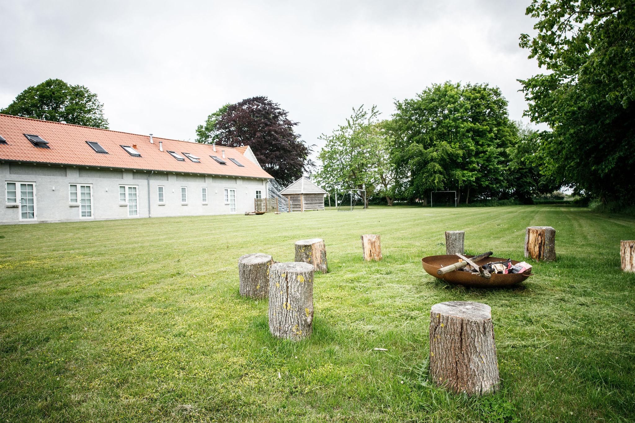 Rund ildsted fyldt med brænde og en økse står på en stor græsplæne, omgivet af en cirkel af træstubbe som siddepladser; i baggrunden ses en lang hvid bygning med rødt tegltag og store træer.