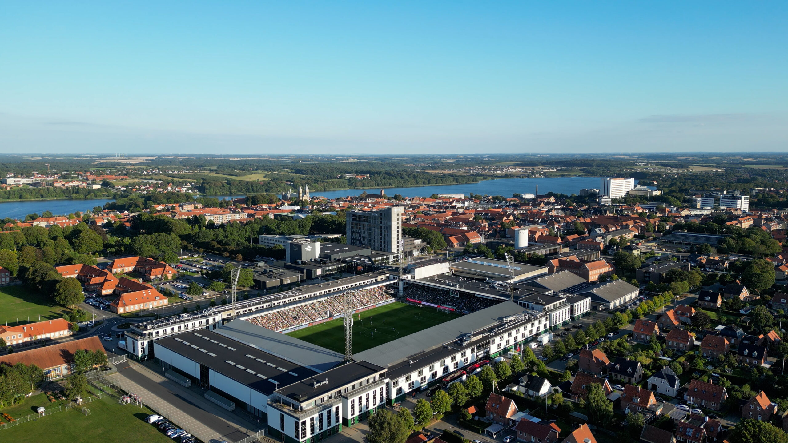 Luftfoto af et stort fodboldstadion med tilskuere på tribunerne midt i en by, omgivet af røde tage, træer og en sø under en klar blå himmel.