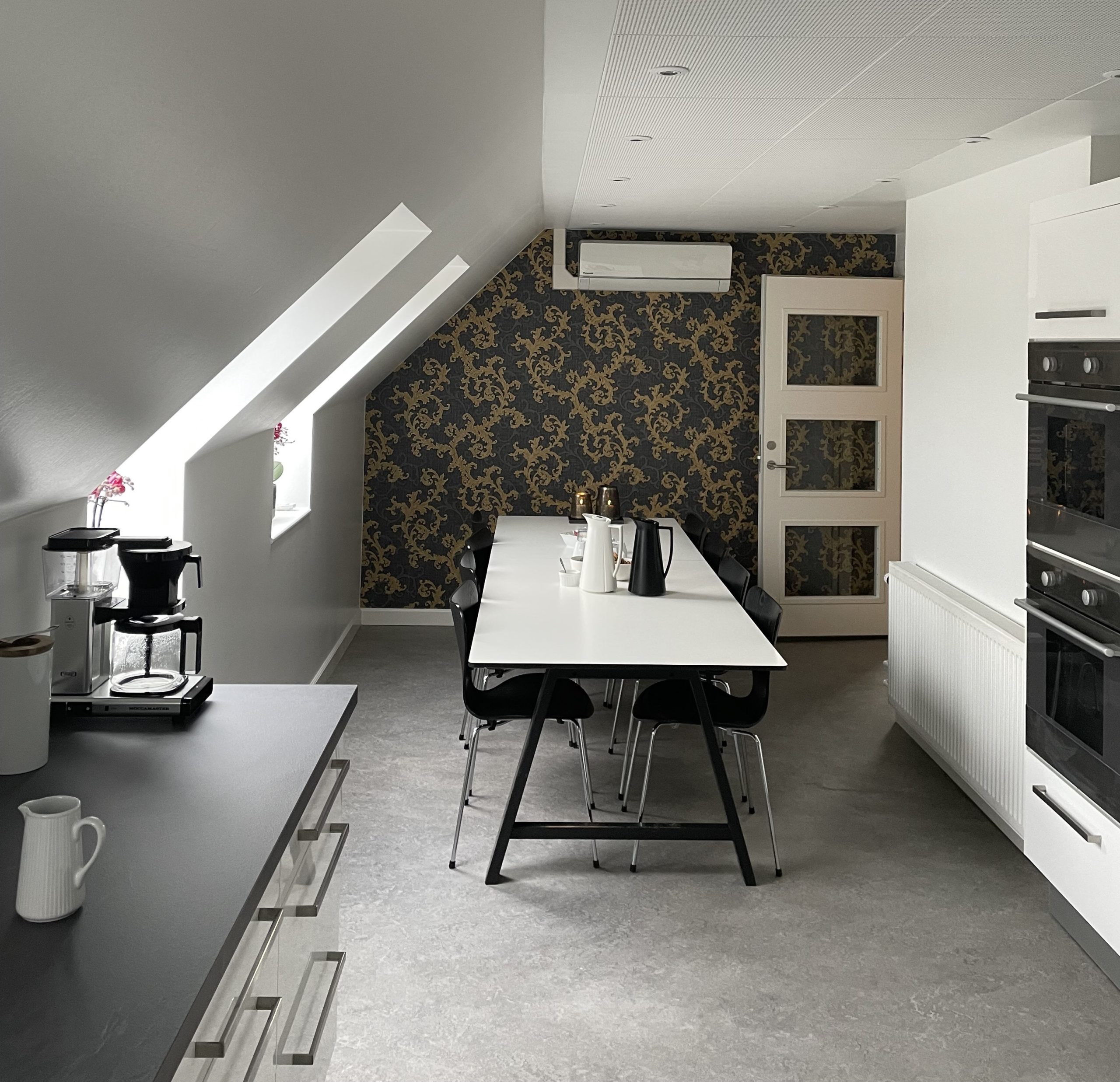 Langt hvidt bord med seks sorte stole står midt i et lofts-køkken og har termokander og kopper på sig; bagvæggen er beklædt med mørkt tapet med gyldent mønster og en dør under en vægmonteret aircondition. Til venstre på en sort bordplade står en kaffemaskine under skrå tagvinduer, og til højre ses indbyggede ovne og en hvid radiator.