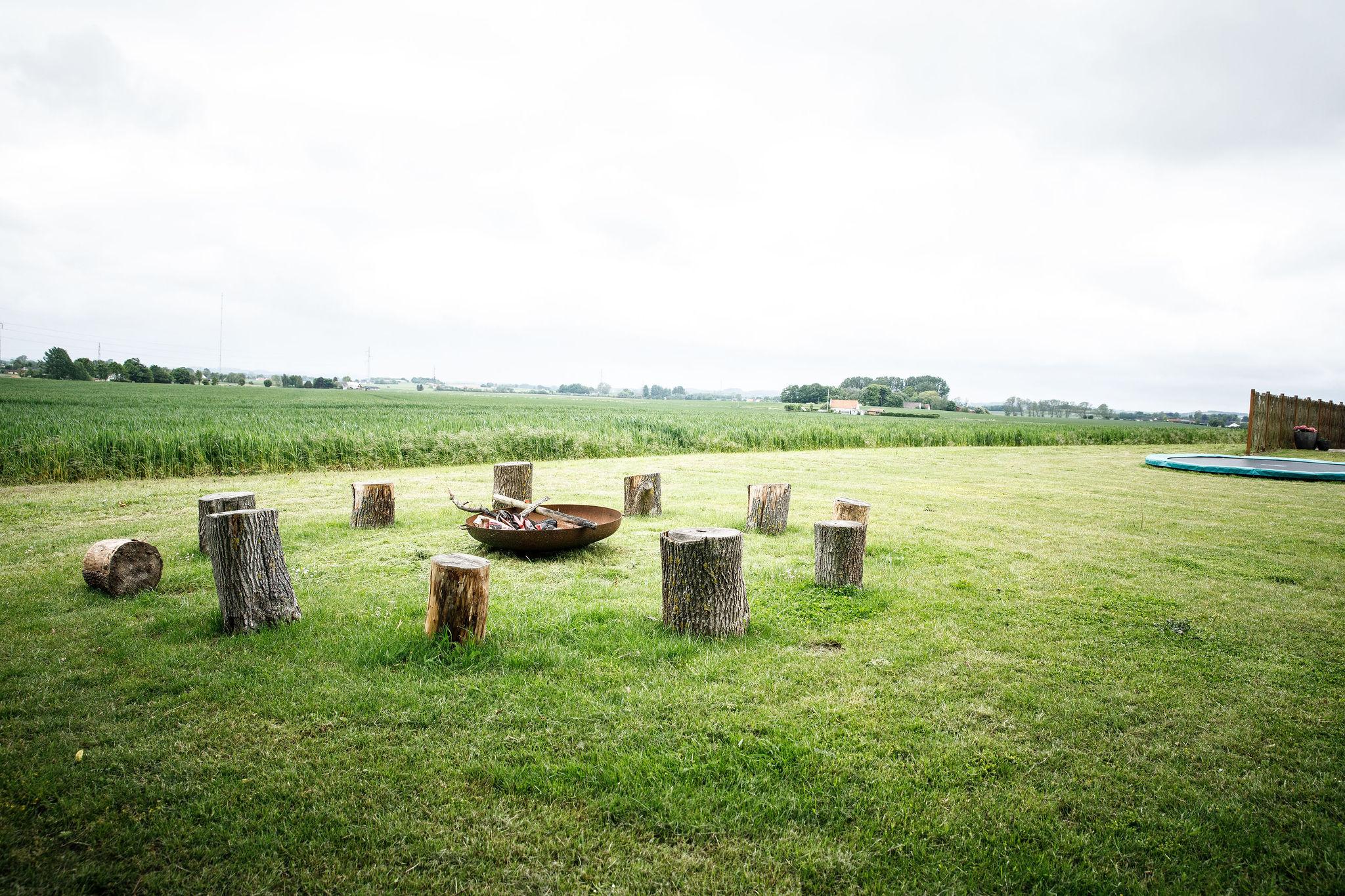 En cirkel af savet træstammer fungerer som siddepladser omkring en rusten ildskål med brænde, placeret på en stor græsplæne med udsigt over åbne marker under en overskyet himmel. En trampolin og et træhegn ses i højre side af billedet.