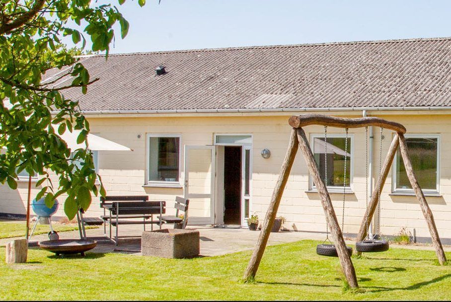 Trægyngestativ med to sorte dækgynger hængende i kæder står på en solbeskinnet græsplæne foran en lav, cremefarvet bygning med åben dør; til venstre ses en terrasse med bord, bænk, parasol og en plante i urtepotte.
