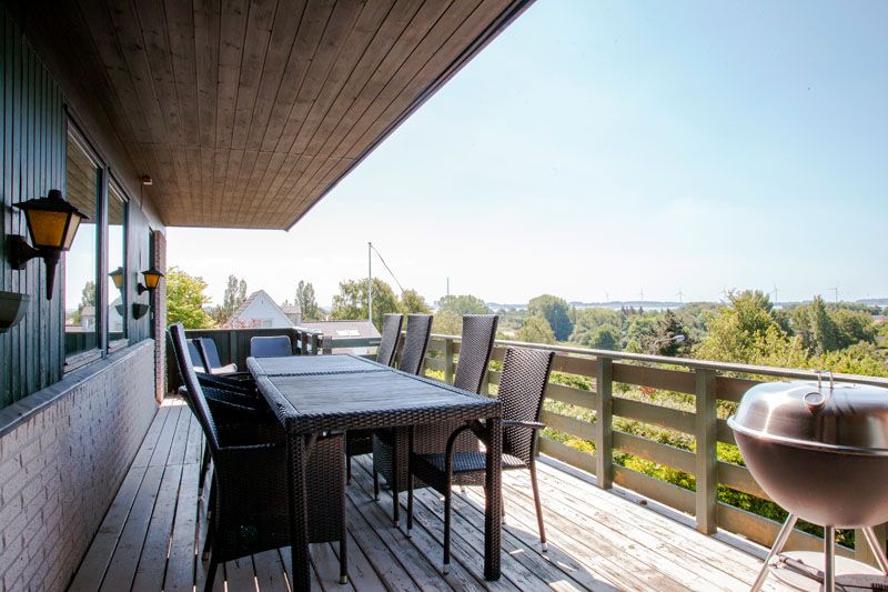 Træterrasse med et langt mørkt fletbord og seks matchende stole under et træbeklædt udhæng. Udsigt over grønne træer og huse i horisonten, og en rund kuglegrill står ved gelænderet til højre.