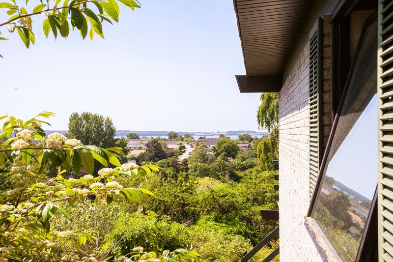 Husets facade med et åbent, skråt vindue til højre, der reflekterer landskabet; forgrunden viser grønne buske og grene med hvide blomster. I midten ses et boligområde og træer, og i baggrunden skimtes kystlinje og hav under en klar blå himmel.