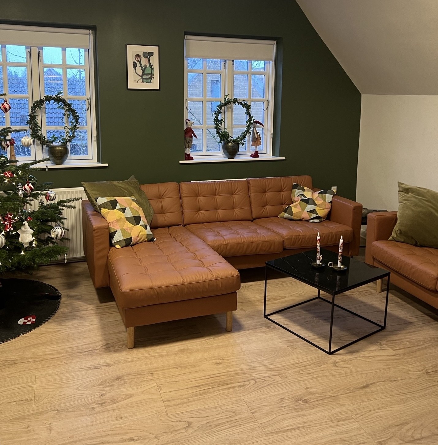 Brun lædersofa i L-form står mod en mørkegrøn væg under to vinduer med julekranse, og et pyntet juletræ ses til venstre. Et sort firkantet sofabord med to dekorerede stearinlys står foran sofaen på lyst trægulv, og mønstrede puder ligger på sofaen.