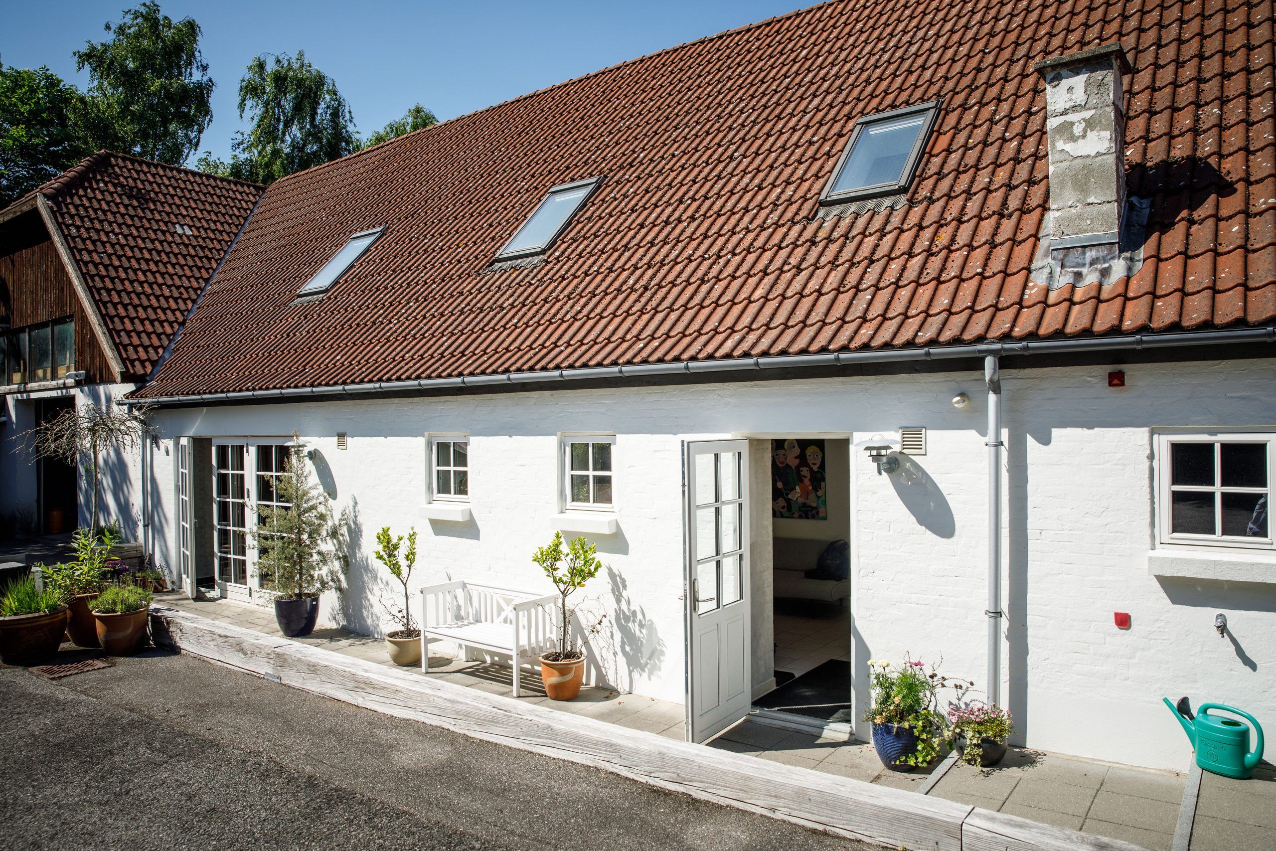 Hvid bygning med rødt tegltag og tre tagvinduer samt en skorsten, hvor en dobbeltdør står åben til et lyst rum. Foran huset er en hvid bænk, flere krukker med planter og en grøn vandkande på en flisebelagt terrasse i solskin.