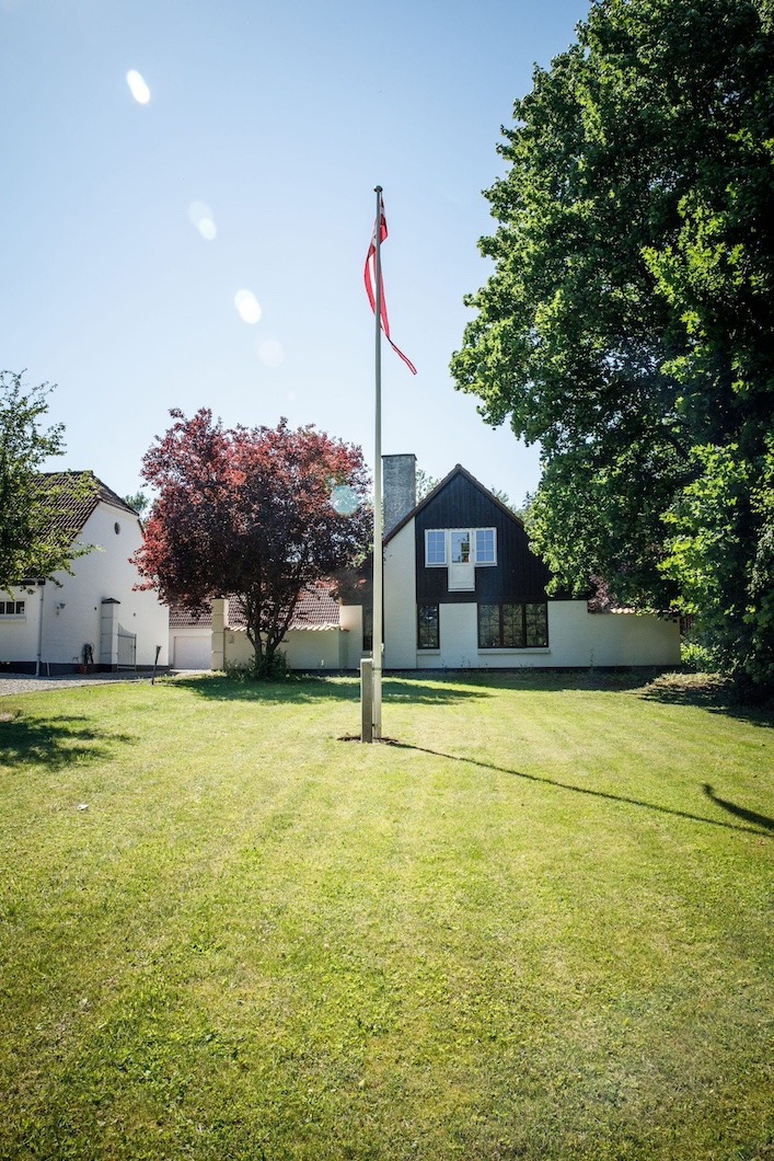 Flagstang med et let svajende dansk flag i midten af en solrig forhave med lysreflekser på himlen. I baggrunden ses et hvidt parcelhus med mørk gavl omgivet af store træer og en veltrimmet græsplæne.