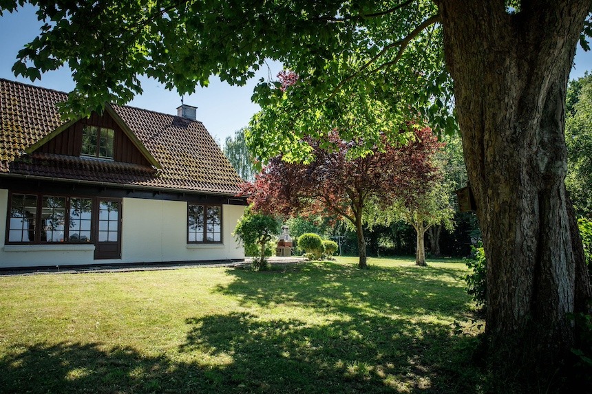 Et hvidt hus med brunt tegltag står til venstre i en solbeskinnet have omgivet af grønne og rødbladede træer. Et stort træ i forgrunden kaster skygge over græsplænen, og en udendørs grill eller pejseovn kan skimtes på en lille terrasse midt i haven.
