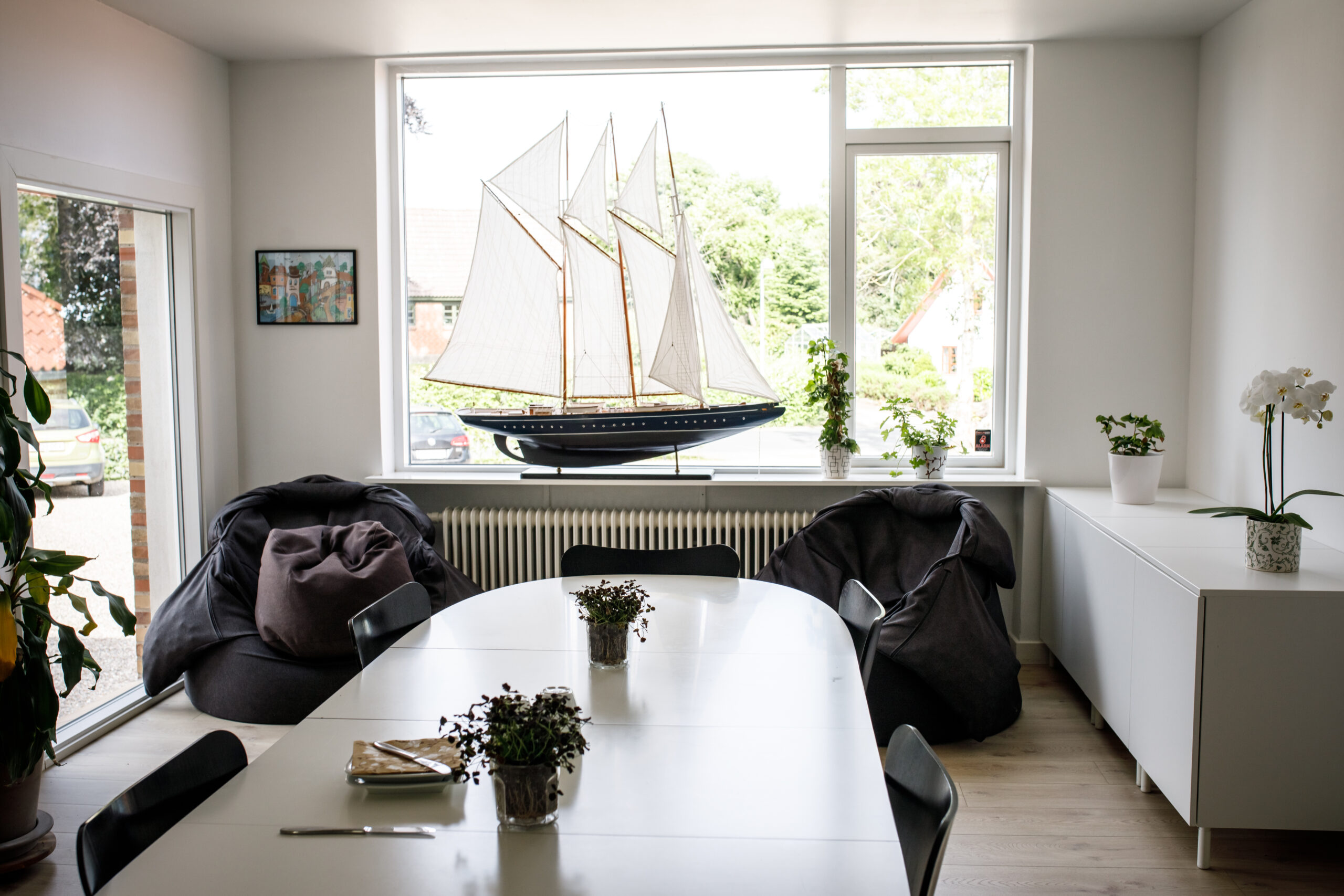 Hvidt ovalt spisebord midt i et lyst rum med små potteplanter og bestik på bordet foran et stort vindue. På vindueskarmen står en stor modelsejlskib, og rummet har to mørke sækkestole, en hvid skænk med en orkidé og udsigt til grønt udenfor.
